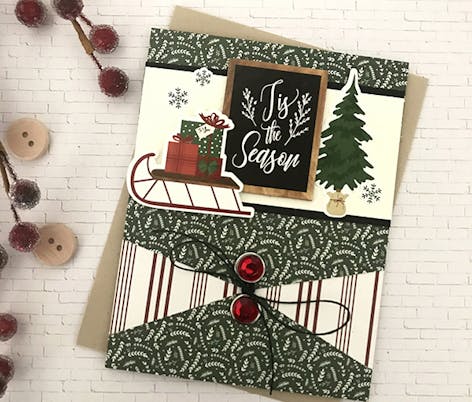 "tis the season" christmas card with small sleight of presents and a tree as well as a envelope-like fold with a ribbon tie