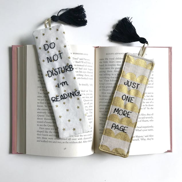 Two fabric bookmarks embroidered in black thread, resting on an open book. One reads 'Do Not Disturb, I'm reading', the second one reads 'Just one more page'